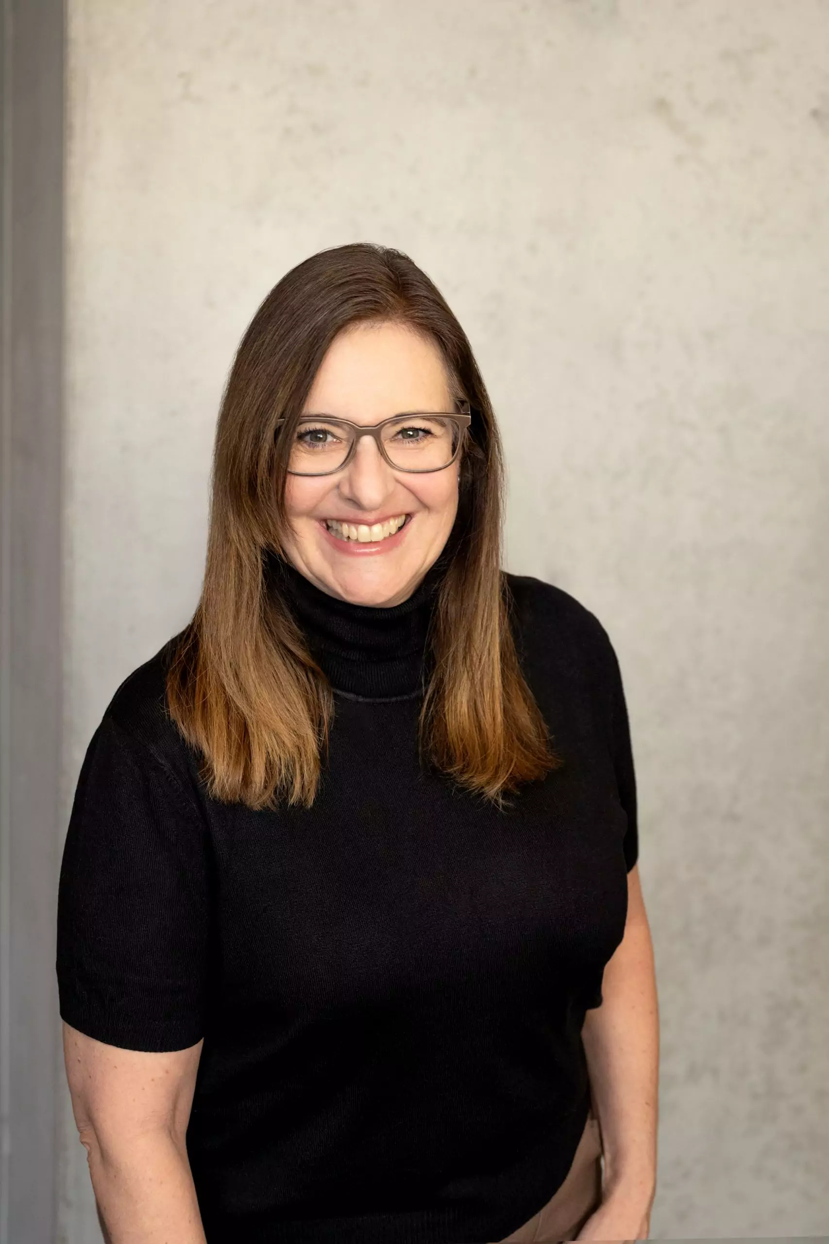 Das Bild ist ein professionelles Porträtfoto einer Frau mit mittel langen, glatten braunen Haaren. Sie hat ein freundliches Lächeln und trägt ein schwarzes Rollkragenshirt. Ihre Arme liegen locker nach unten in Richtung Hüfte. Sie trägt eine Brille, in einem schlichten hellbraunen Design. Der Hintergrund ist eine schlichte, leicht strukturierte, graue Wand. Die Beleuchtung ist weich und natürlich, wodurch ihr Gesicht gut zur Geltung kommt. Die gesamte Bildkomposition wirkt professionell und eignet sich gut für geschäftliche oder berufliche Zwecke.