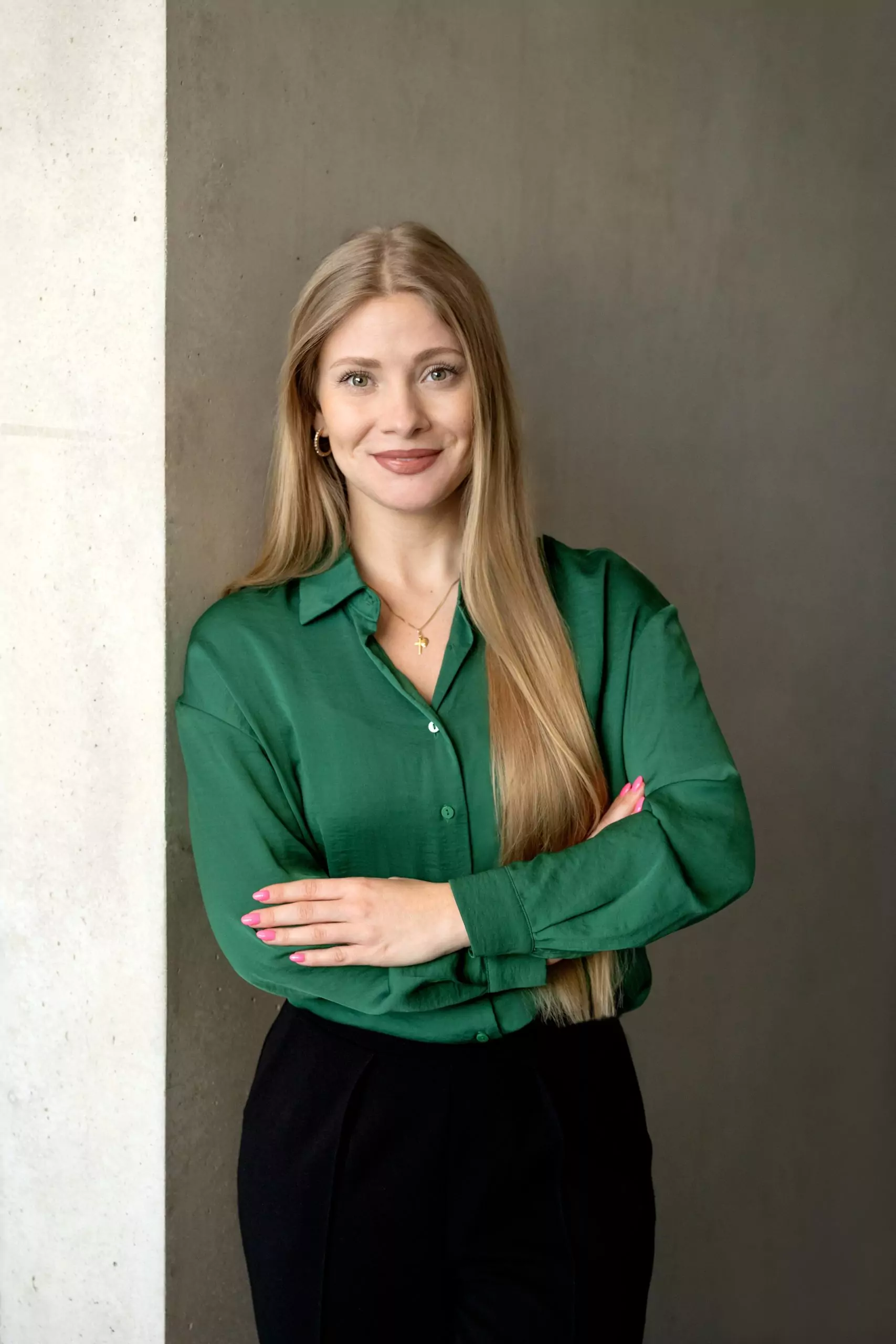 Das Bild ist ein professionelles Porträtfoto einer Frau mit langen, glatten blonden Haaren. Sie hat ein freundliches Lächeln und trägt einen grüne Bluse. Ihre Arme sind verschränkt, und sie trägt eine goldene feine Kette. Der Hintergrund ist eine schlichte, leicht strukturierte, graue Wand. Die Beleuchtung ist weich und natürlich, wodurch ihr Gesicht gut zur Geltung kommt. Die gesamte Bildkomposition wirkt professionell und eignet sich gut für geschäftliche oder berufliche Zwecke.