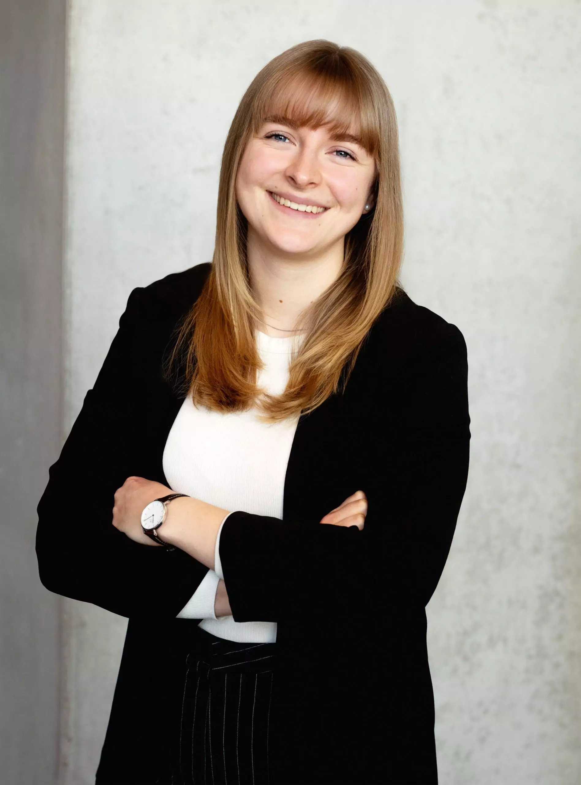 Das Bild ist ein professionelles Porträtfoto einer Frau mit mittellangen, glatten dunkelblonden Haaren. Sie hat ein freundliches Lächeln und trägt einen schwarzen Blazer über einem weißem Oberteil. Ihre Arme sind verschränkt und sie trägt eine schwarz goldene Uhr mit einem schlichten Design an ihrem linken Handgelenk. Der Hintergrund ist eine schlichte, leicht strukturierte, graue Wand. Die Beleuchtung ist weich und natürlich, wodurch ihr Gesicht gut zur Geltung kommt. Die gesamte Bildkomposition wirkt professionell und eignet sich gut für geschäftliche oder berufliche Zwecke.